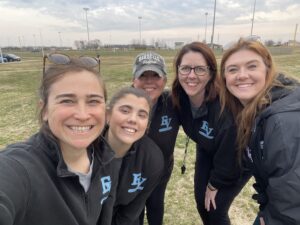 Five women smiling and leaning together for a selfie on a grassy sports field. They are wearing matching black jackets with light blue “EV” logos, suggesting they are part of the same team or staff. One woman is wearing a gray baseball cap, and two have sunglasses resting on their heads. A softball or baseball complex with fences, light poles, and a cloudy sky appears in the background.