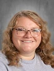 Professional headshot of a smiling woman with glasses and wavy blonde hair against a grey background.
