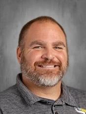 A headshot of a smiling man with a beard, wearing a grey polo shirt against a textured grey background.
