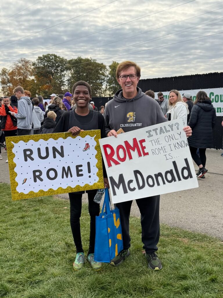 Rome McDonald poses with coach Mike Radtke for a photo. Rome is holding a sign that reads "Run Rome!" Mike Radtke is holding a sign that says "Italy? The only Rome I know is Rome McDonald."