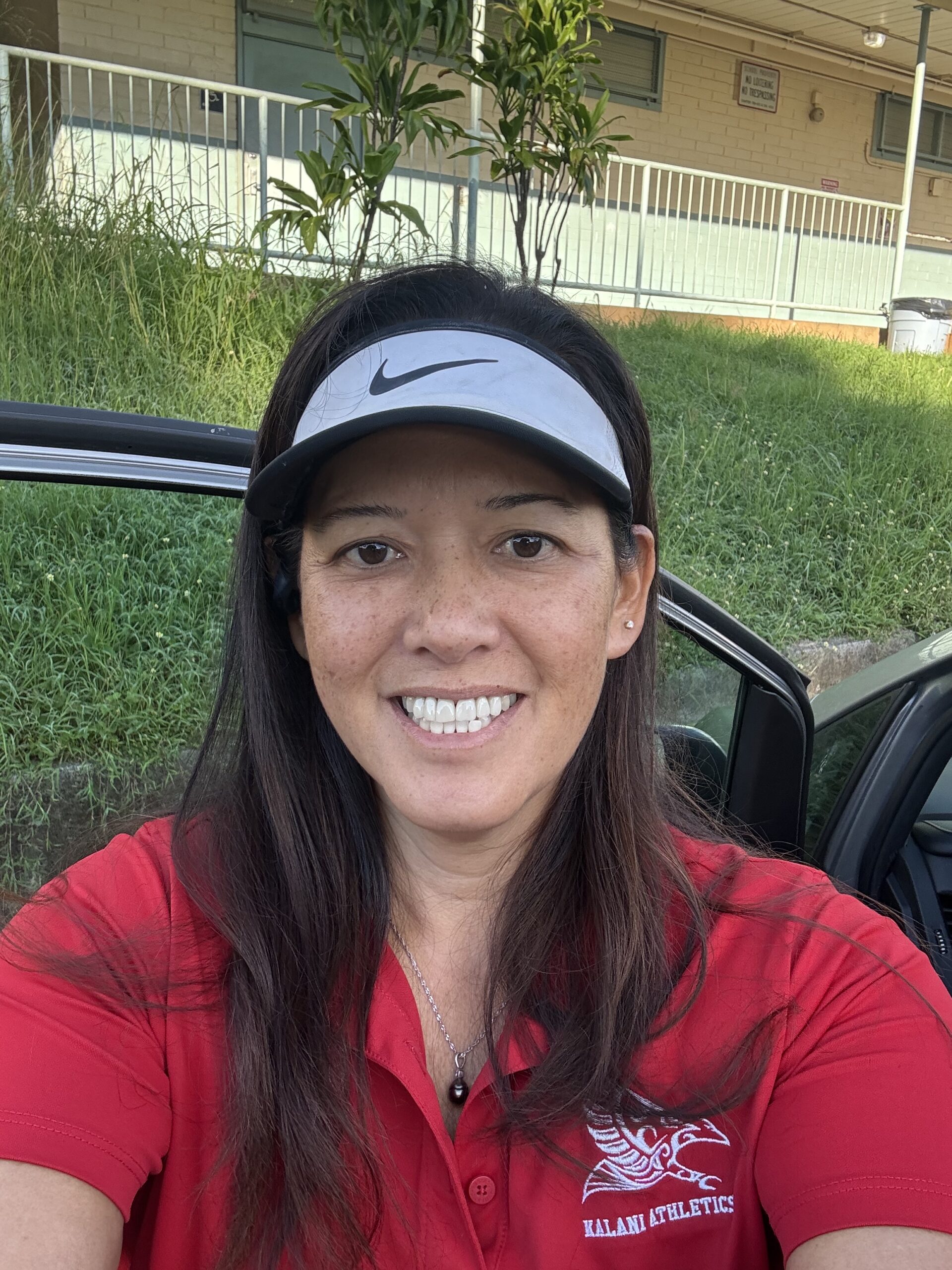Headshot of a smiling woman with long dark hair and a gray Nike visor, wearing a red polo shirt with a "Kalani Athletics" logo.