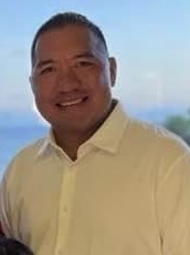 Headshot of a smiling man with short dark hair, wearing a white collared shirt, with a blurred outdoor background.