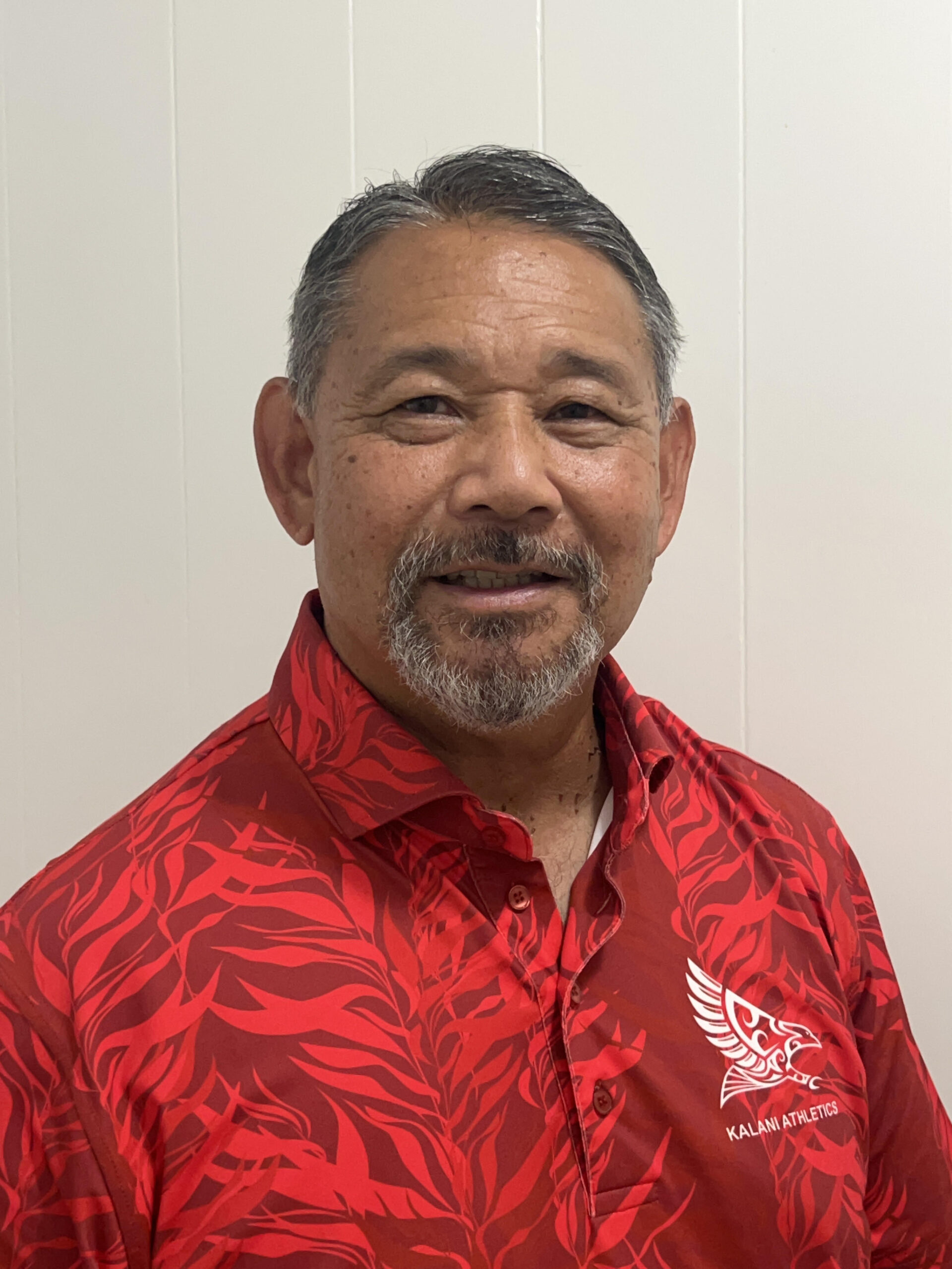 Headshot of a smiling man with a gray beard wearing a red short-sleeved collared shirt with a "Kalani Athletics" logo.