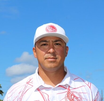 Headshot of Coach Kenric Pai wearing a white hat and a white polo shirt with red trim, standing outdoors under a blue sky.