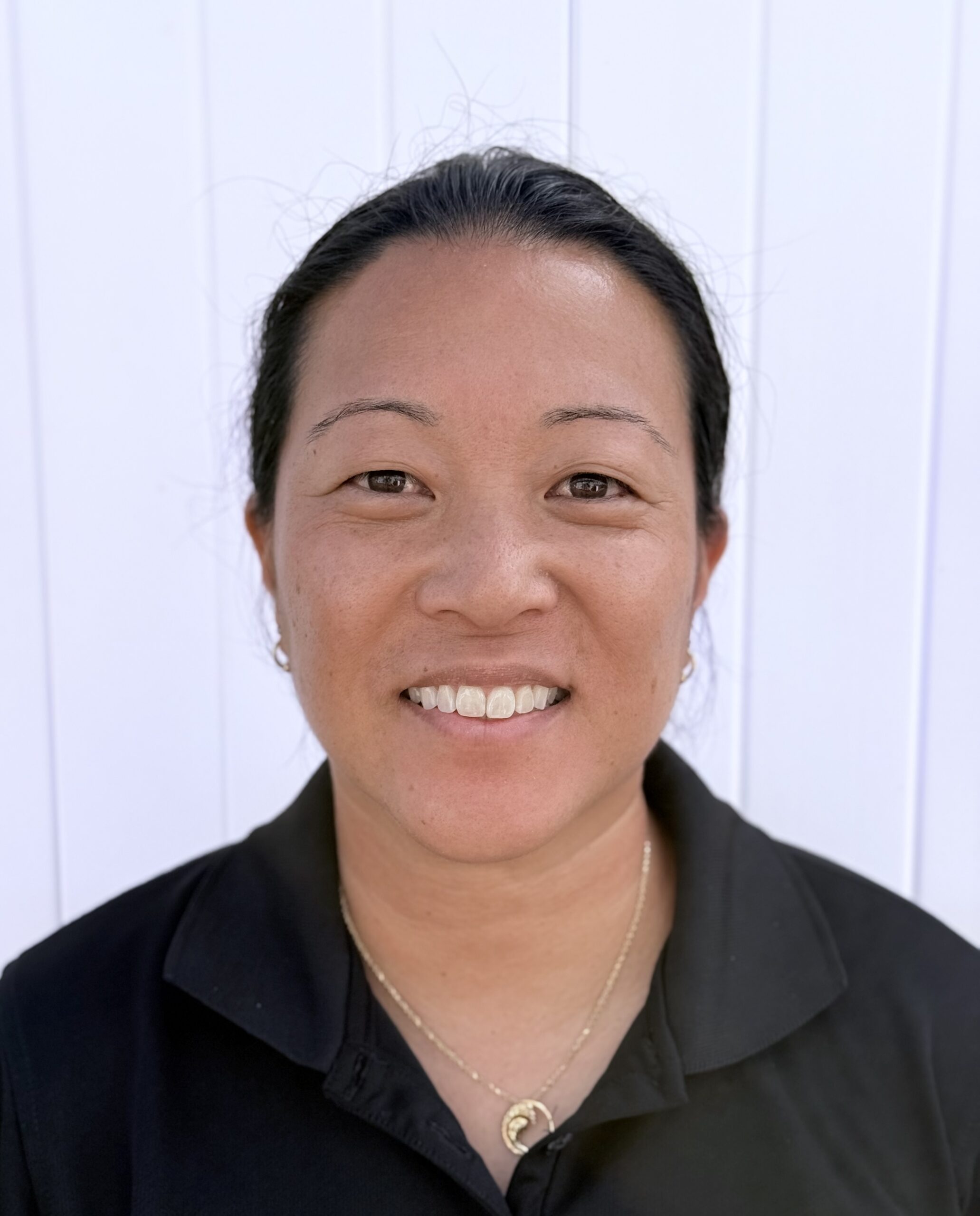 Headshot of a woman, Coach Jillian Okamoto, smiling and wearing a black collared shirt and a gold necklace, standing in front of a white wall.