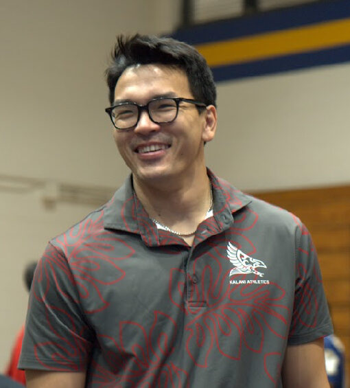Coach Jason Tran smiling indoors, wearing glasses and a gray polo shirt with red designs and a Kalani Athletics logo.