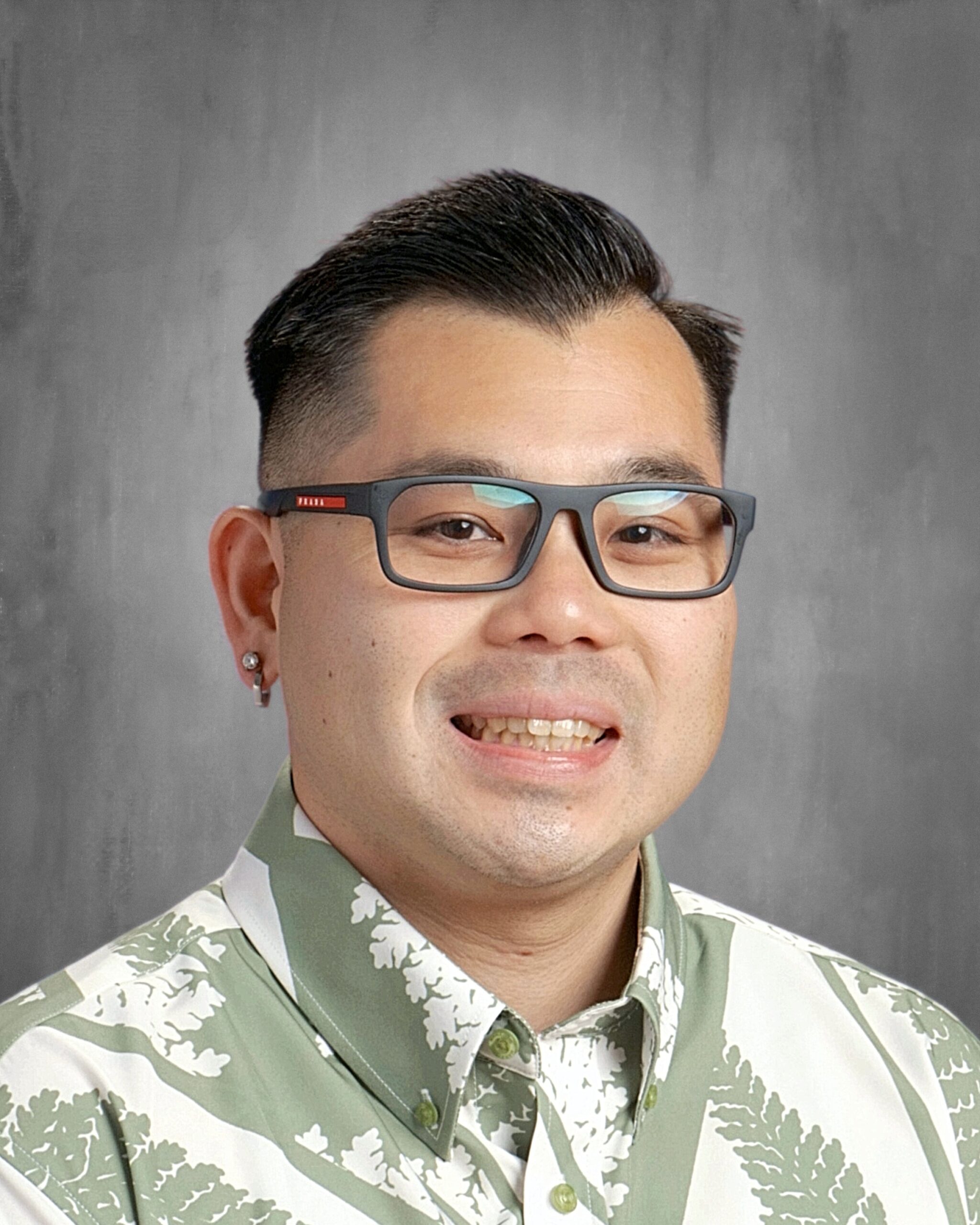 Headshot of a smiling man in glasses, wearing a light green and white patterned aloha shirt against a gray background