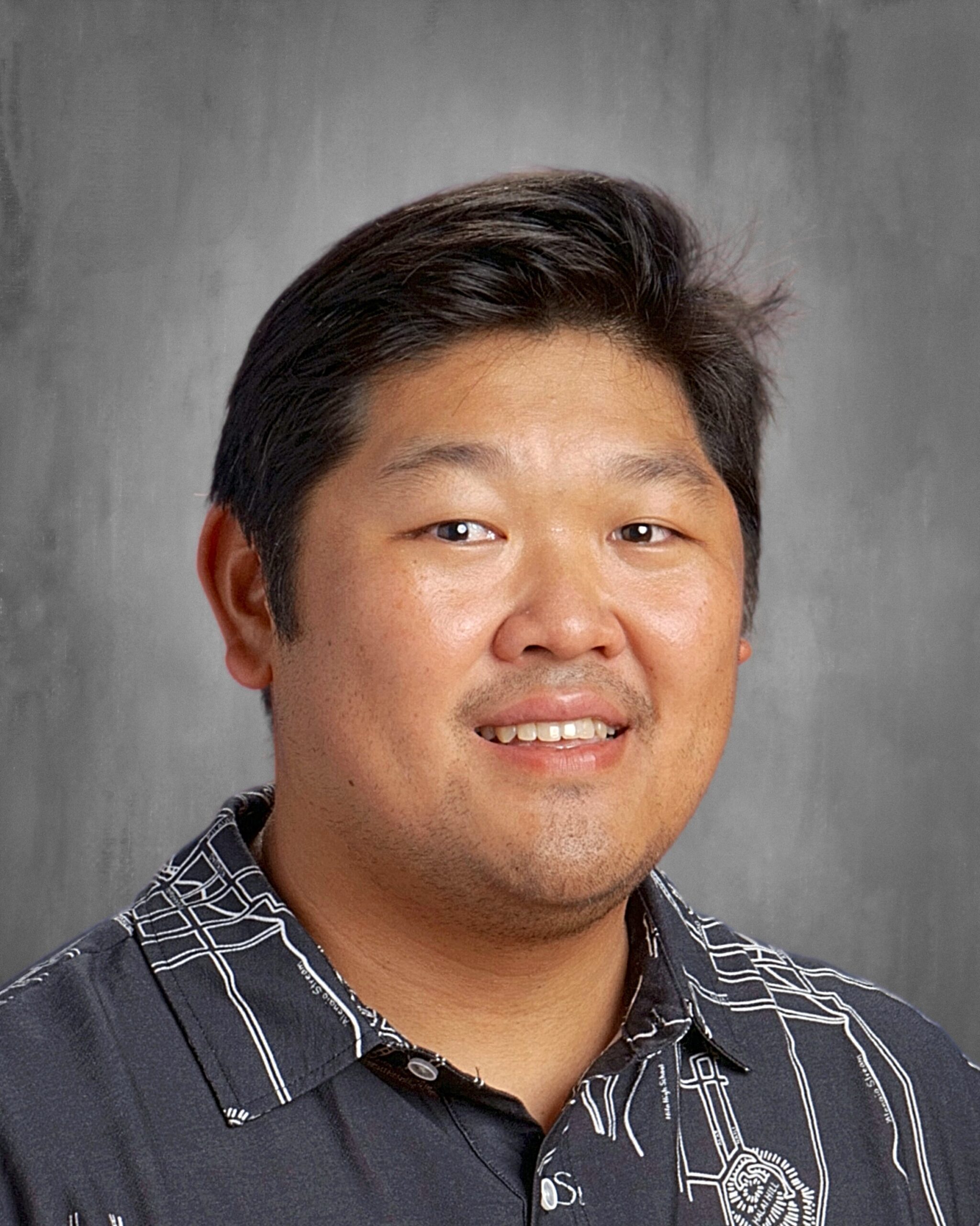 Headshot of a smiling man with dark hair, wearing a dark patterned aloha shirt against a gray background