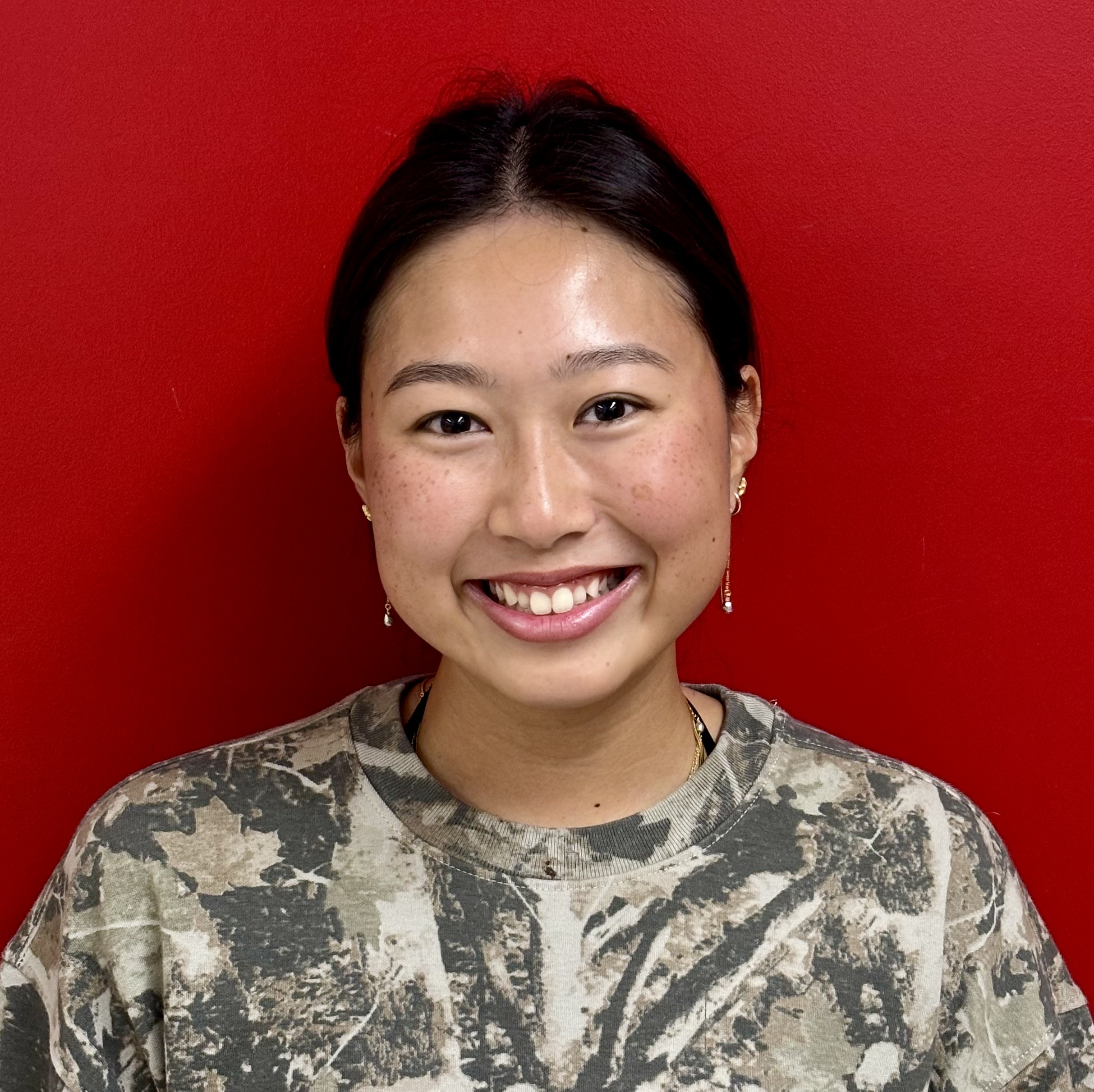 Headshot of a smiling young woman with dark hair tied back, wearing a camouflage shirt against a red background.
