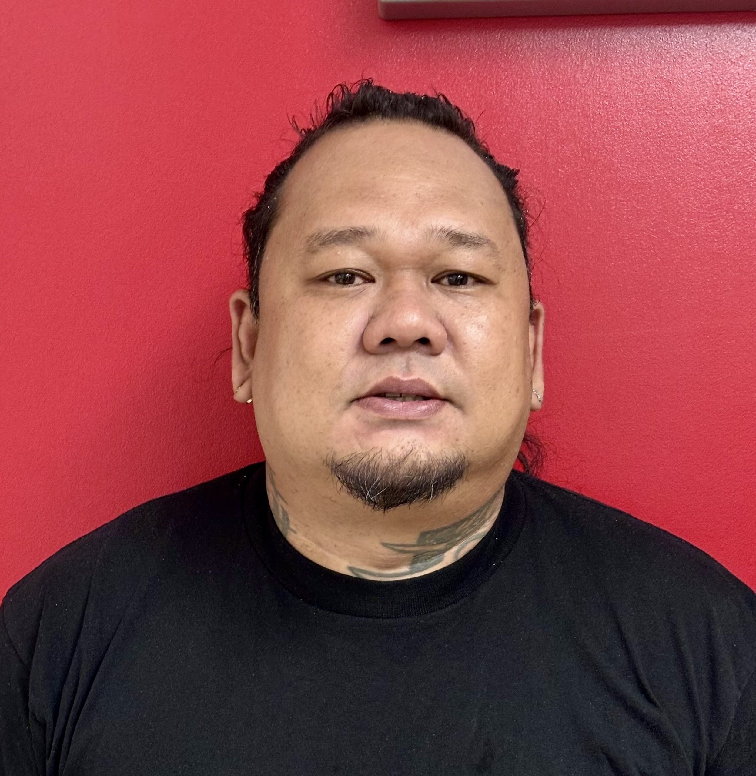 Headshot of a man with dark hair tied back, a goatee, and neck tattoos, wearing a black t-shirt against a red background.