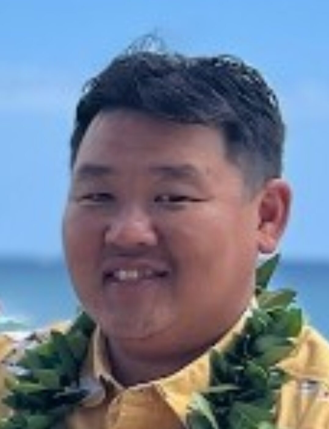 Headshot of a smiling man with short dark hair, wearing a yellow Hawaiian shirt and a green lei, with a blurred beach background