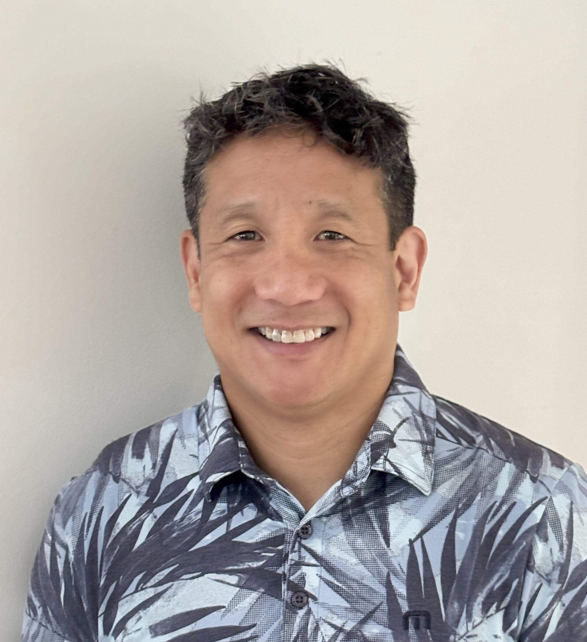 Headshot of a smiling man with short, dark curly hair, wearing a blue and black patterned aloha shirt