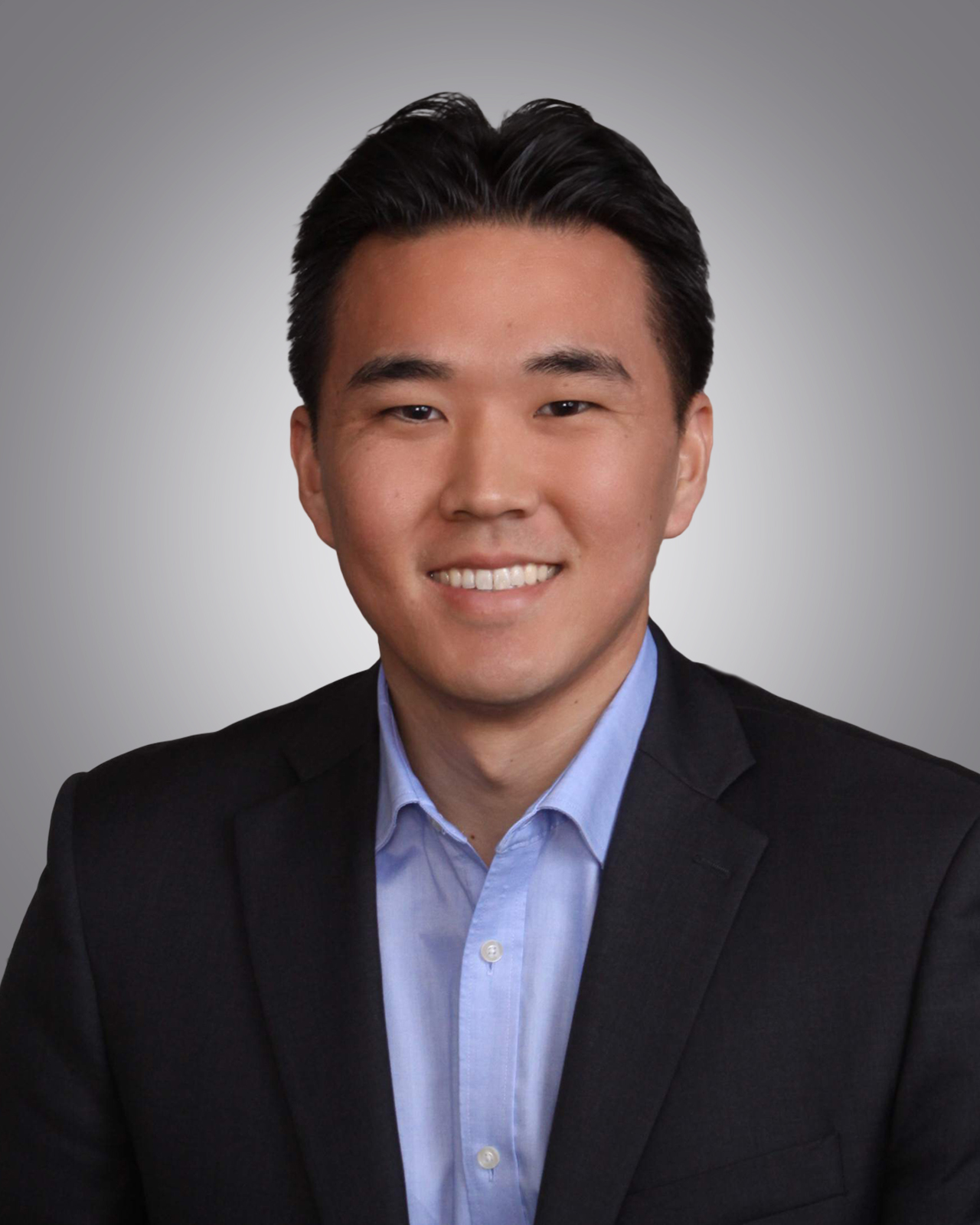 Headshot of a smiling young man with dark hair, wearing a black suit jacket and a light blue collared shirt
