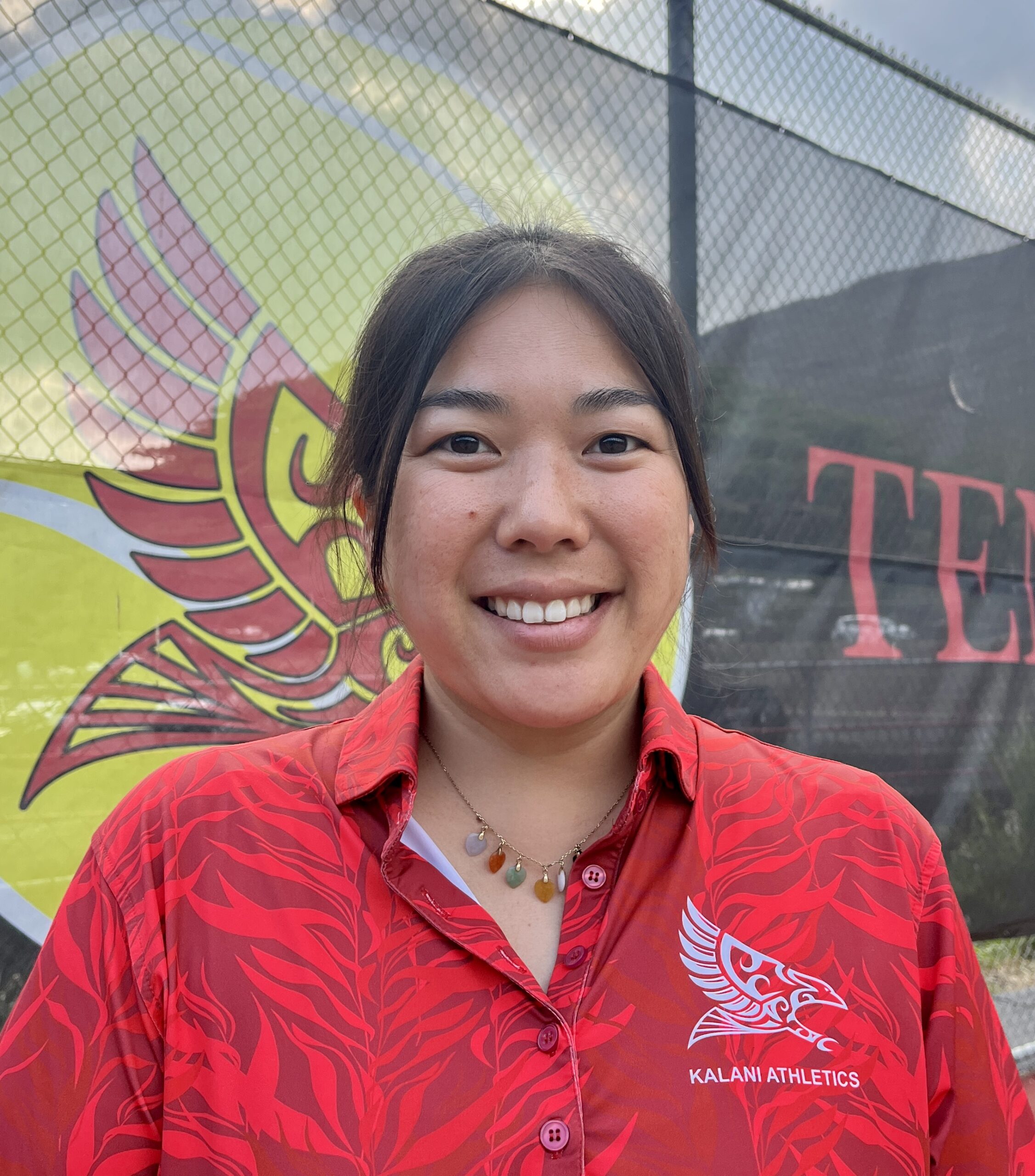 Headshot of a smiling young woman with dark hair, wearing a red patterned polo shirt with a "Kalani Athletics" logo. A yellow and black banner with a bird design is visible in the background.