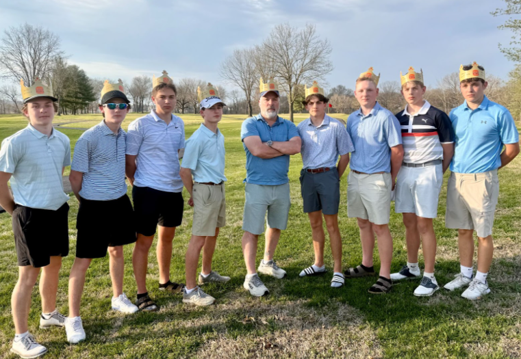 Nine young men standing in a line in the grass at a Golf Course. All are wearing shorts and polos and burger kind crowns.