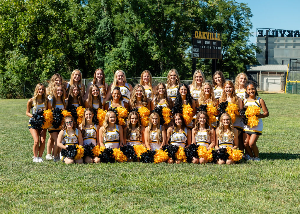 Three rows of cheerleaders in white, yellow, and black cheer uniforms with black and yellow poms in a green grass firld.