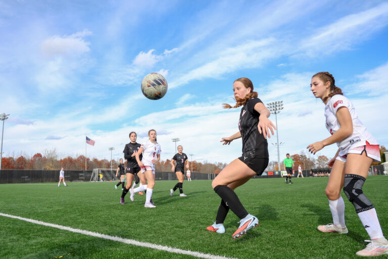 A girls soccer player in a navy jersey tries to volley the ball out of the air with a defender behind her.