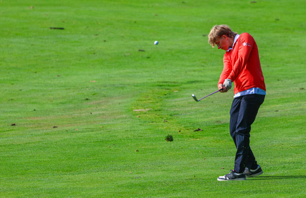 A golfer in a red shirt and black pants hits a ball and with a divot flying in the photo.