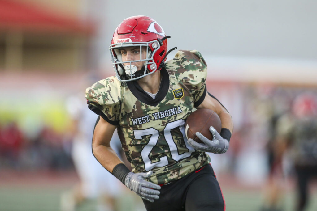 A player with a red helmet and camaflouge jersey runs with the football.