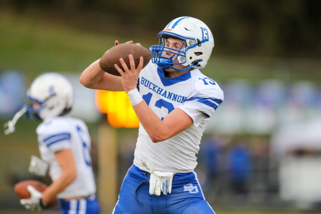 A Buckhannon-Upshur football player wearing No. 13 in a white jersey and blue pants catches a football during pregame warmups, with a teammate holding a ball in the background.