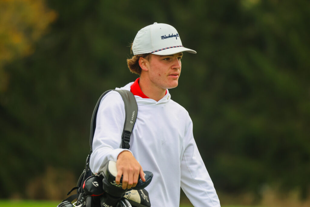 A golfer in a white hooded pullover and light-colored cap walks the course carrying his golf bag over his shoulder, with trees blurred in the background.