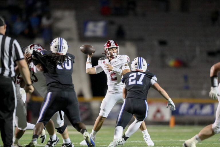 A quarterback in a white No. 2 jersey and maroon helmet sets to throw as a defender in a navy No. 27 jersey rushes toward him during a night football game, with linemen engaged and an official in the foreground.
