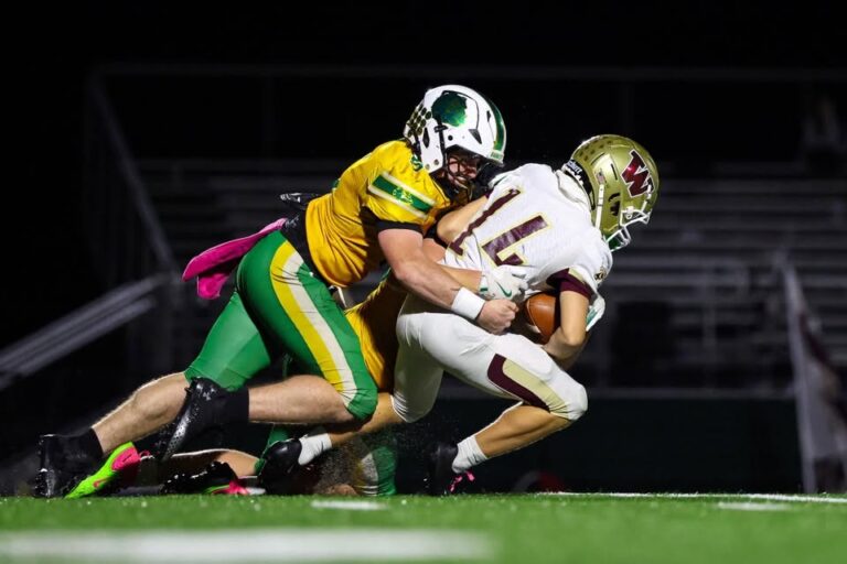 A defender in a gold jersey and green pants wraps up a ball carrier in a white No. 14 jersey and gold helmet during a night football game.