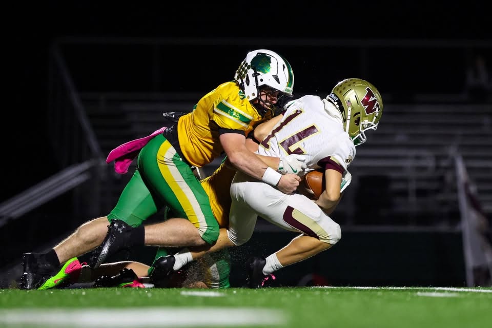 A defender in a gold jersey and green pants wraps up a ball carrier in a white No. 14 jersey and gold helmet during a night football game.
