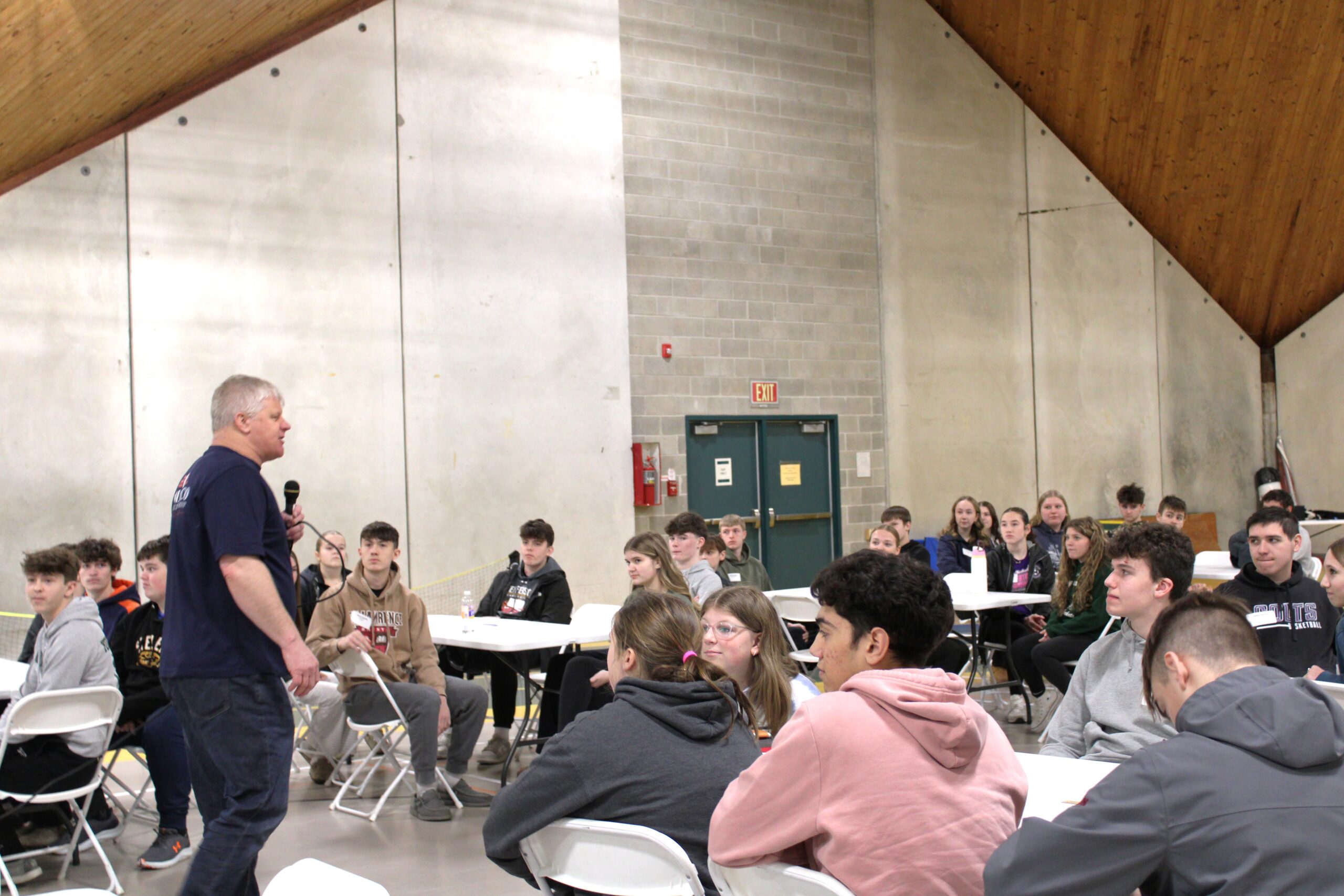 Large group of students seated in chairs and listening to an adult male holding a microphone