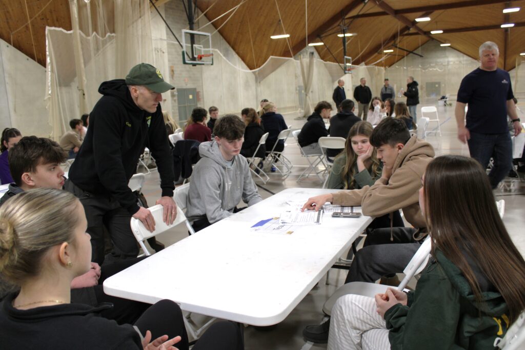 Small group of students sitting around a white table working together on paperwork with an adult male assisting their work