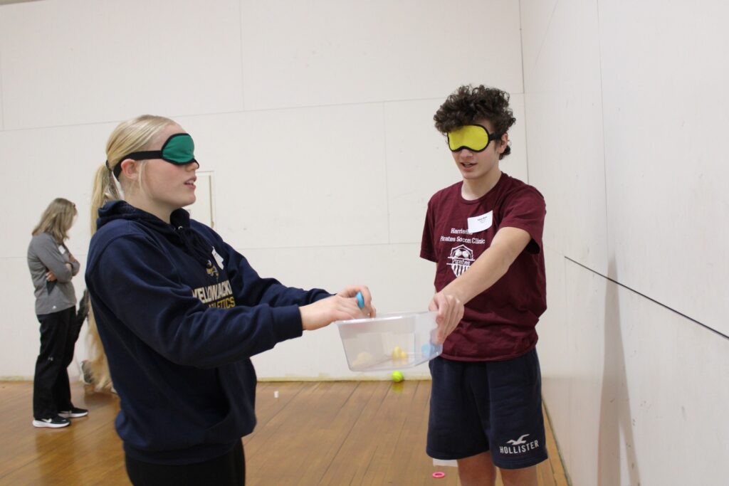 A female and male student wearing blindfolds working together to gather items into a small plastic tub