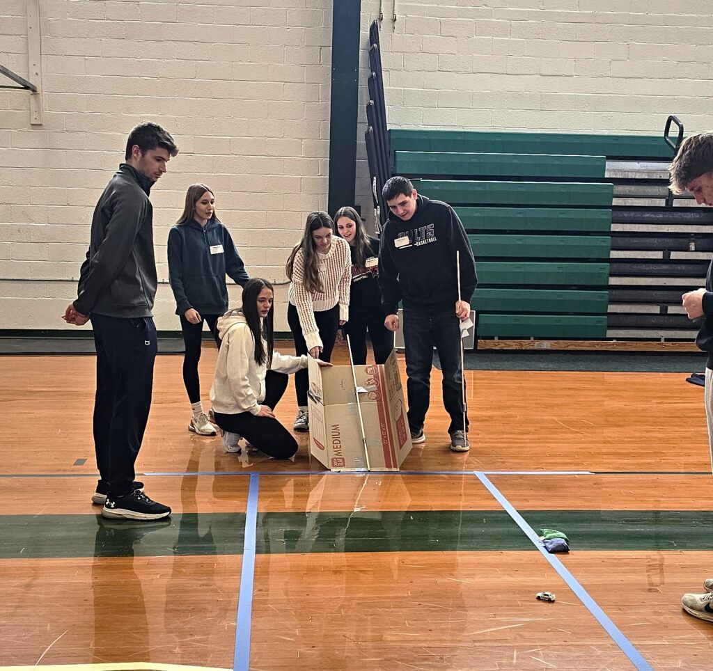 Small group of students standing and kneeling next to a cardboard-like track watching a toy car roll down