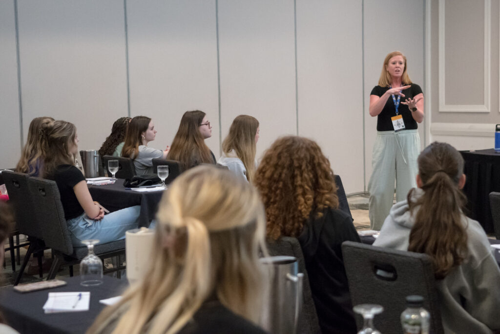 Large group of high school students seated in a conference room listening to an adult female give a presentation