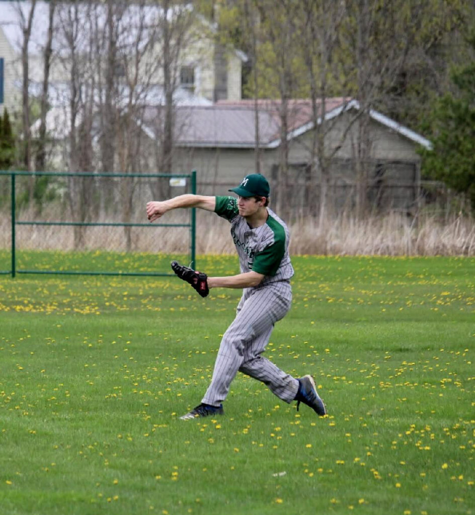 Male high school student wearing a green and white cap and gray and green uniform with a black baseball glove on his left hand and right arm extended