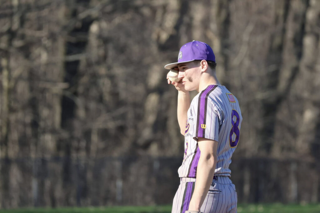 Male high school student wearing a purple cap and a gray and purple uniform holding a baseball
