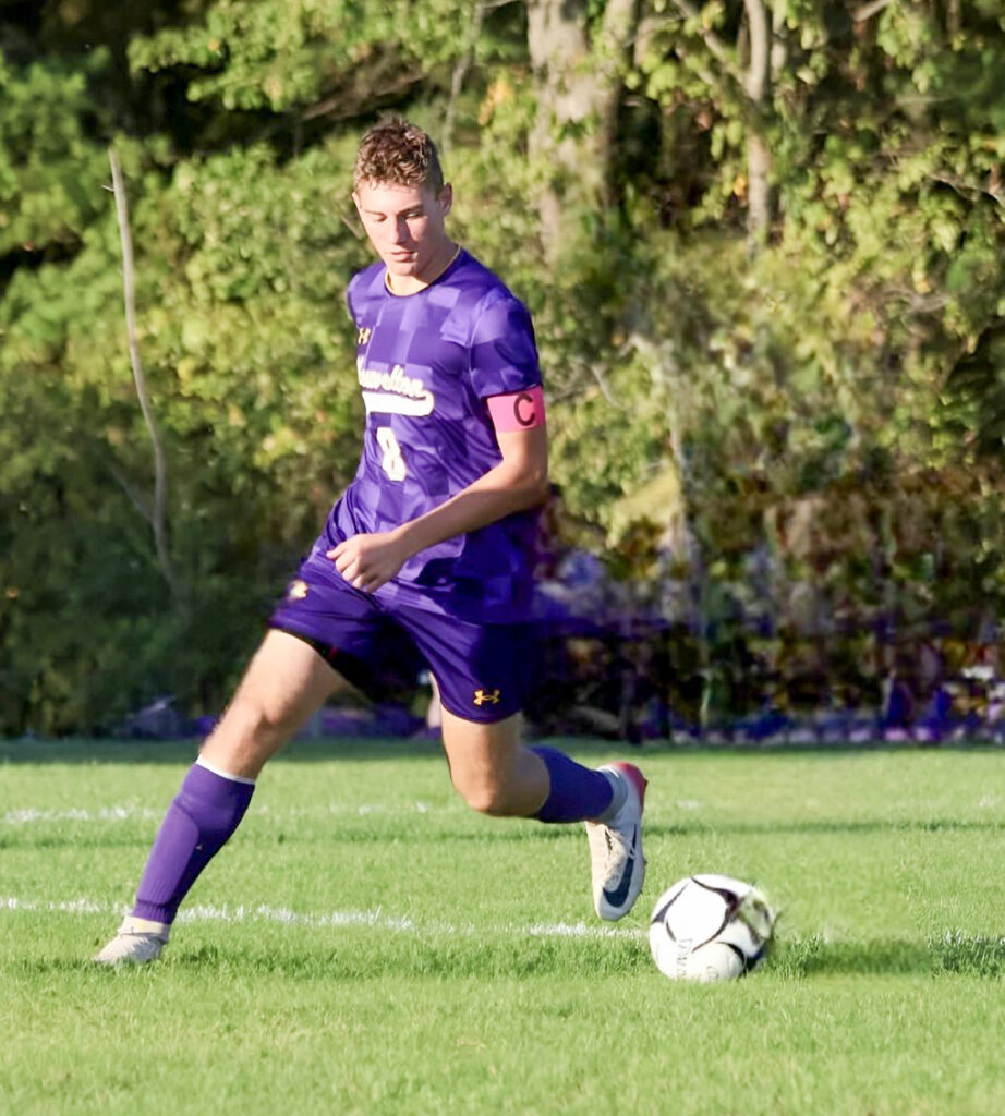 Male high school student wearing a purple uniform and socks with a white and black soccer ball on the grass in front of him