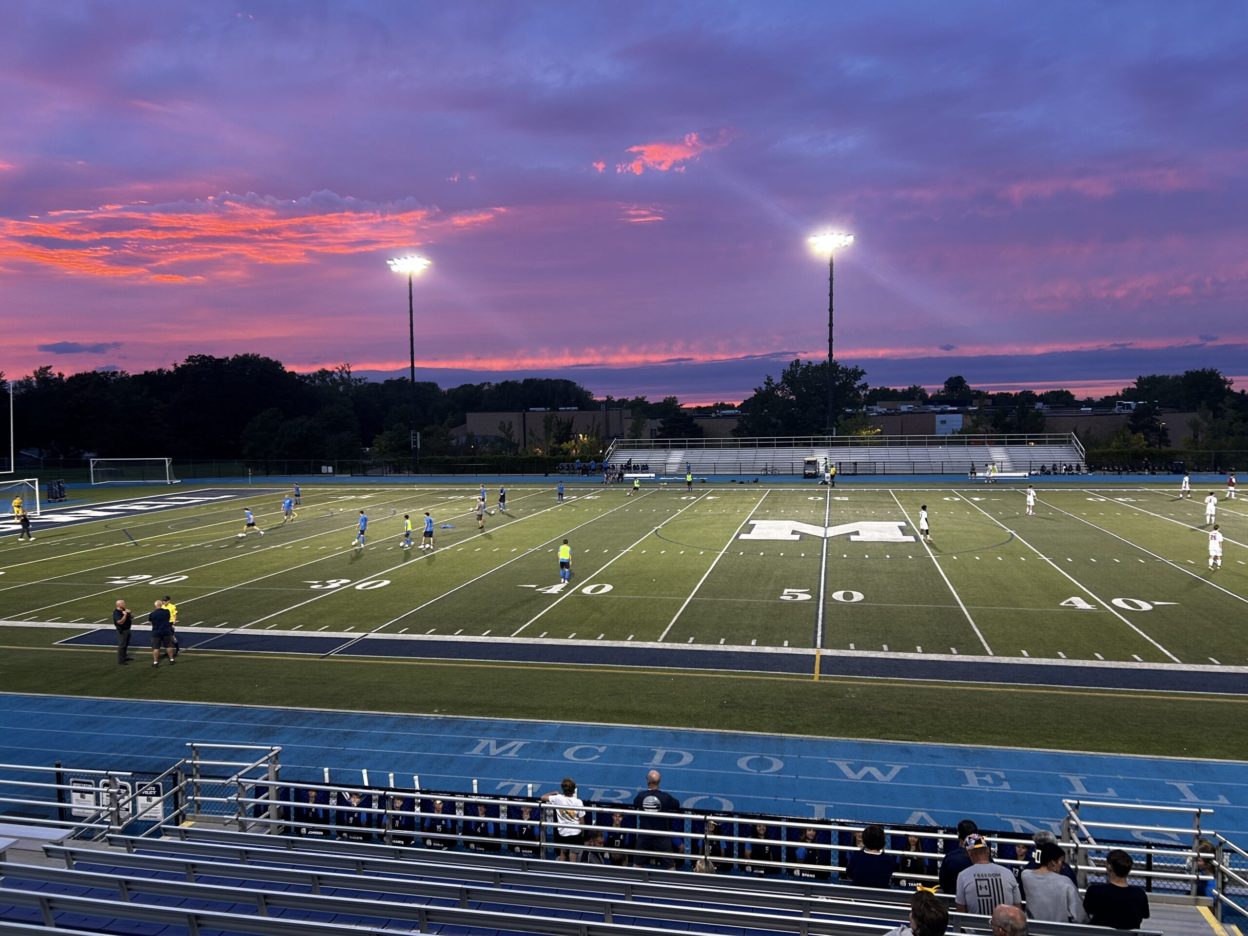 A high-angle, wide shot of a soccer field at night illuminated by stadium floodlights, with a twilight sky in the background.