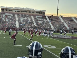 Wide view of a high school football game in an outdoor stadium, showing players lined up on the field with bleachers and green hills in the background.