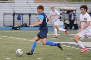 A McDowell High School varsity boys' soccer player in a blue uniform kicking a soccer ball mid-match on an outdoor field.
