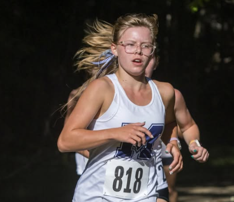 A female McDowell cross country runner wearing a white and navy singlet with bib number 818, glasses, and a headband, running intently during a race.