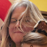 A person with glasses smiles warmly. Hair falls across their face. Next to them, a child wearing leopard ears, partially visible, creates a playful tone.